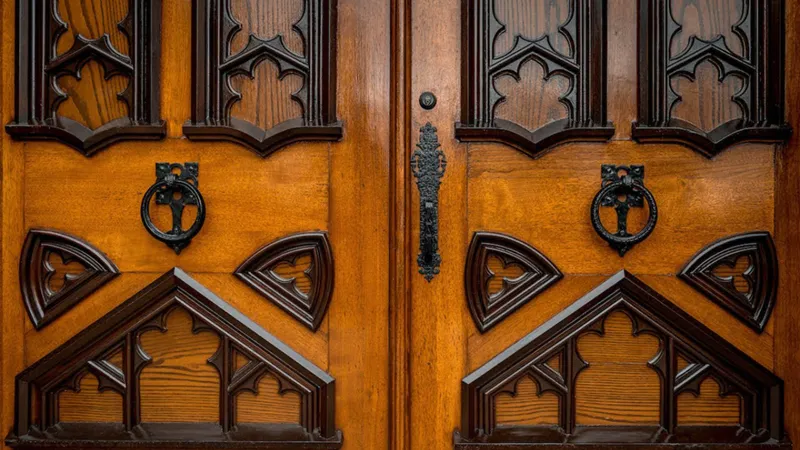 A pair of ornate wooden doors at the University of Notre Dame featuring dark Gothic-style carvings. Symmetrical black metal ring handles are mounted on the doors, which are joined by a decorative black iron latch in the center.